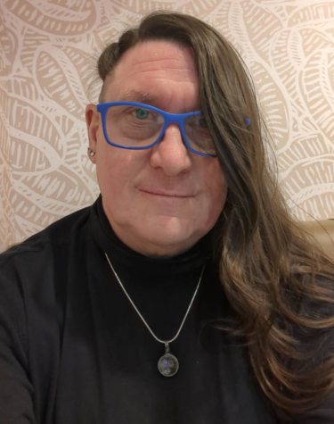 trans woman with long hair and an undercut on the side wearing blue broad rimmed glassed, wearing a silver chain and a black polo neck smiles warmly towards the camera